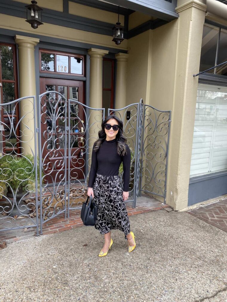 Woman standing with a midi-length skirt featuring white faces printed on a black background and wearing a black turtleneck.