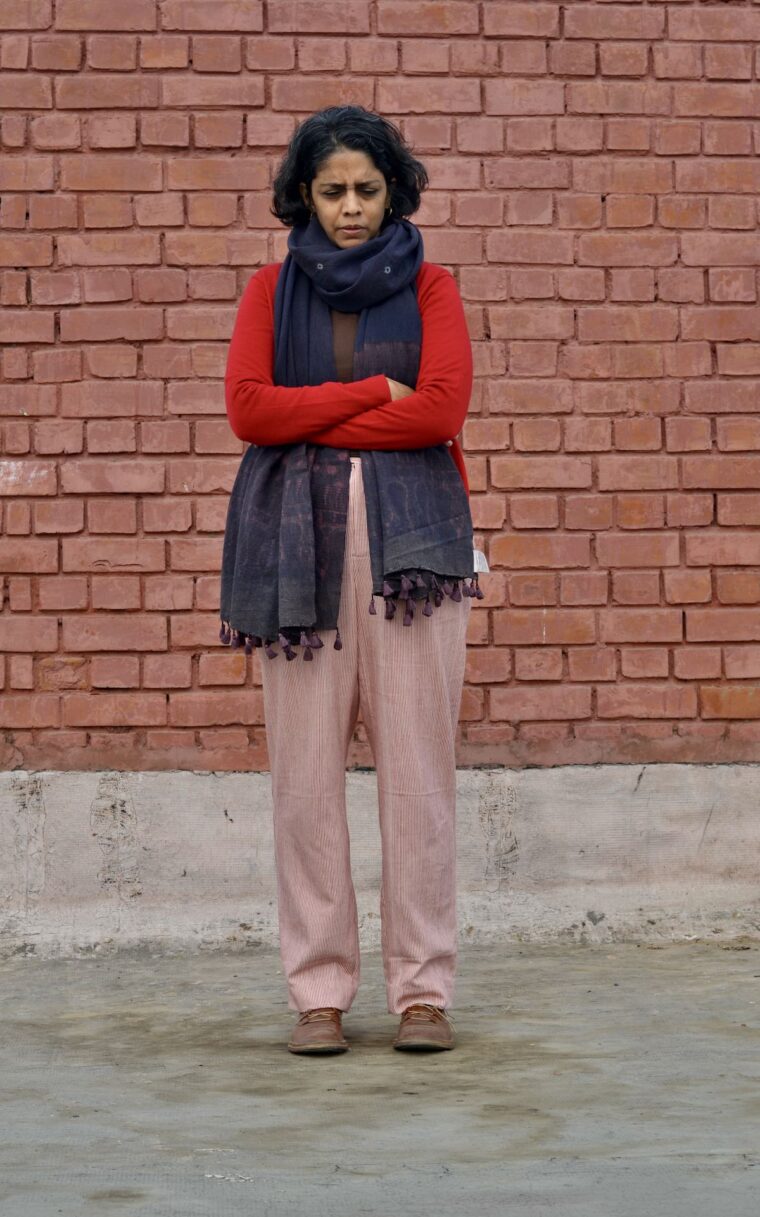 Woman standing in front of a brick wall wearing red and white trousers, a red sweater and long scarf. She is looking at down at the ground in front of her.