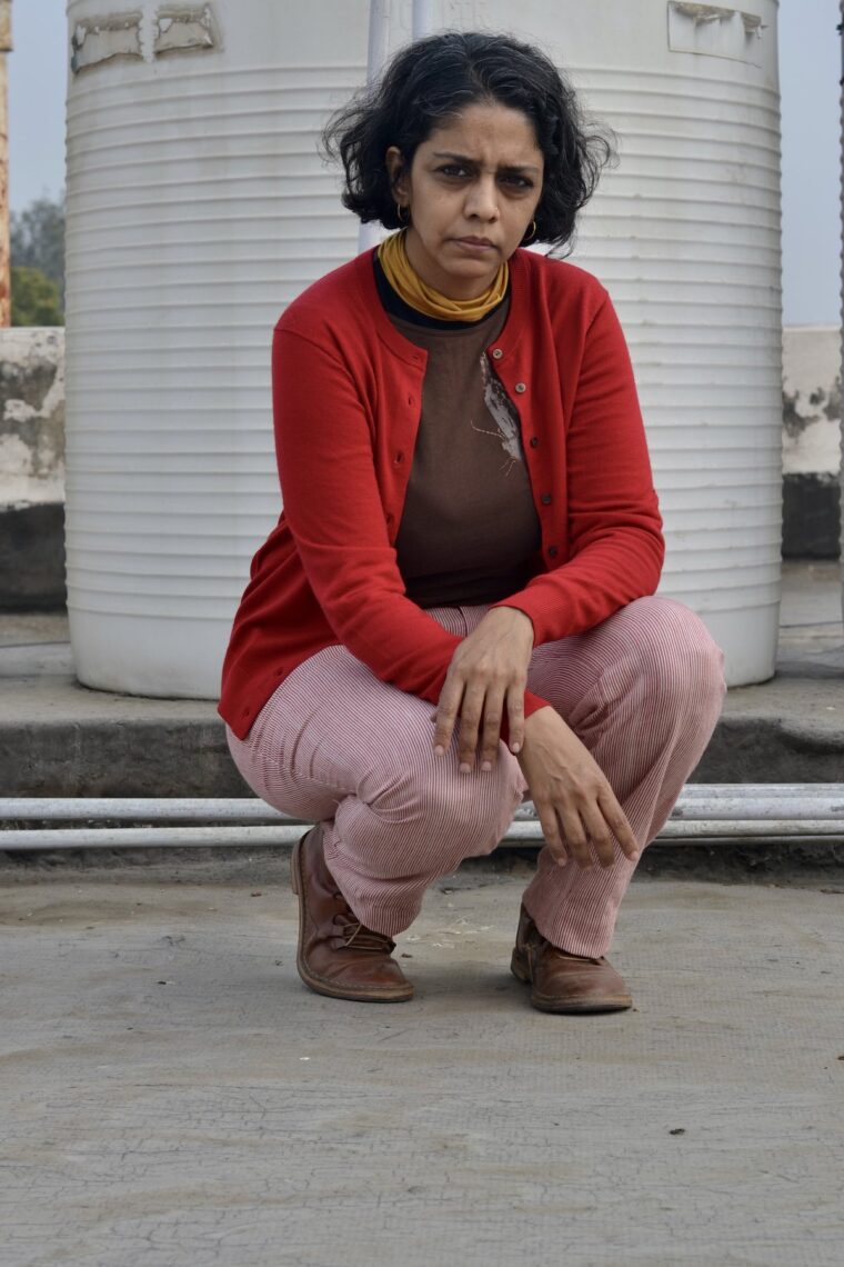 Woman squatting in red and white trousers, wearing a brown sweater and a red cardigan.