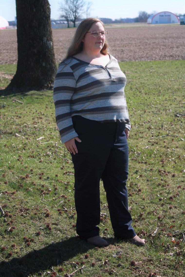 Woman standing outside in a striped knit henley shirt with long sleeves and navy pants.
