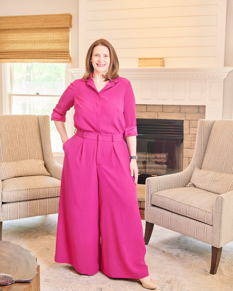 Woman wearing pink wide-legged trousers and matching pink blouse. She is standing in a living room in front of a fire place. She has her hand in a pocket