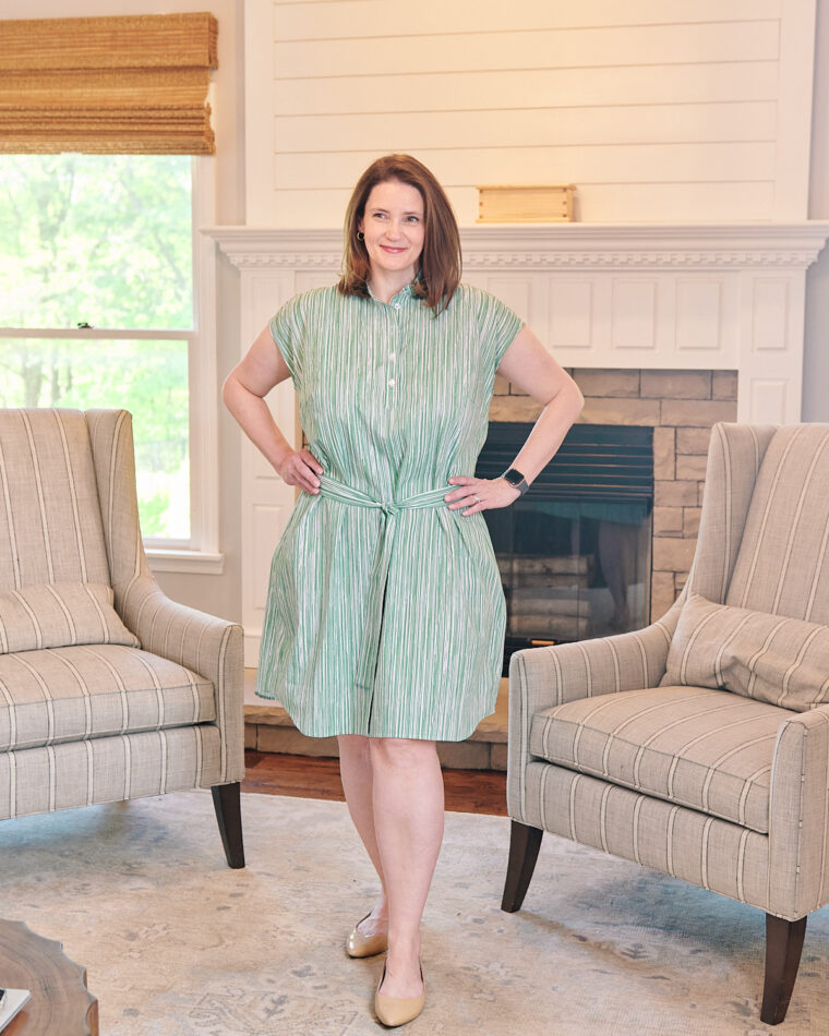 A smiling woman wearing a green shirt dress is standing in a living room in front of a fireplace. She has both hands on her hips. Her dress is belted in a matching fabric.