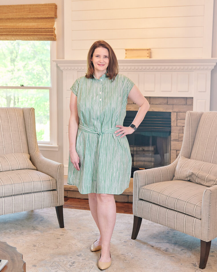 A smiling woman wearing a green shirt dress is standing in a living room in front of a fireplace. She has one hand on her hip. Her dress is belted in a matching fabric.
