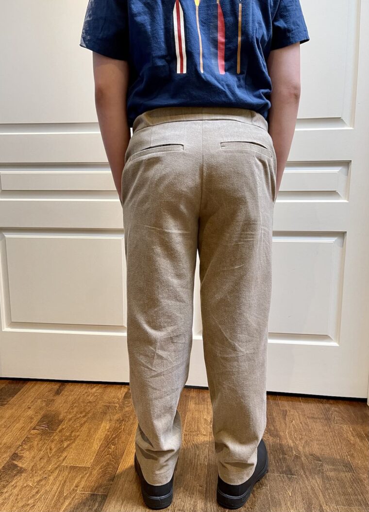 Back view of a young man wearing tan trousers and navy blue top. Picture is from the shoulders down.His hands are in his pocket.