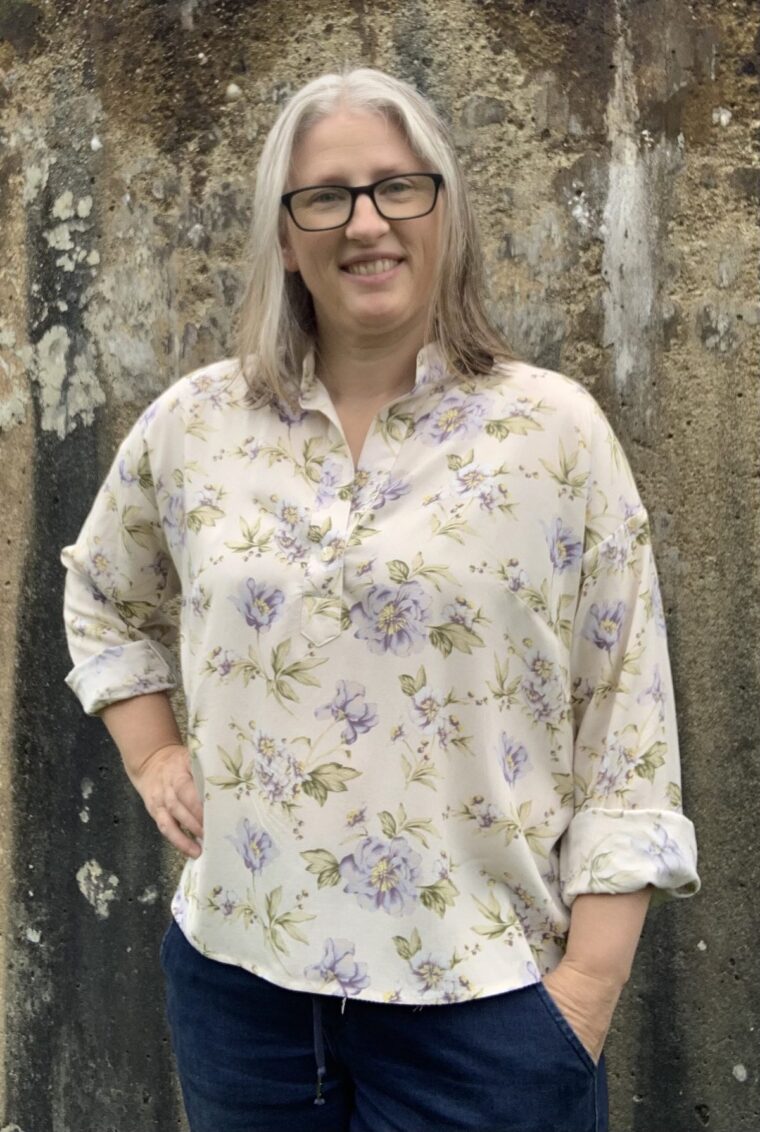 Smiling woman wearing a pullover long-sleeve blouse made in a drapey floral fabric. She has her hand on her hip.