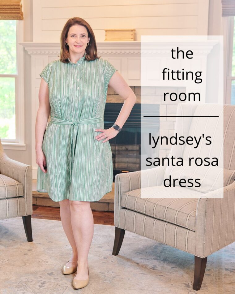 A smiling woman wearing a green shirt dress is standing in a living room in front of a fireplace. She has one hand on her hip. Her dress is belted in a matching fabric. The text on the image says "The Fitting Room: Lyndsey's Santa Rosa Dress"