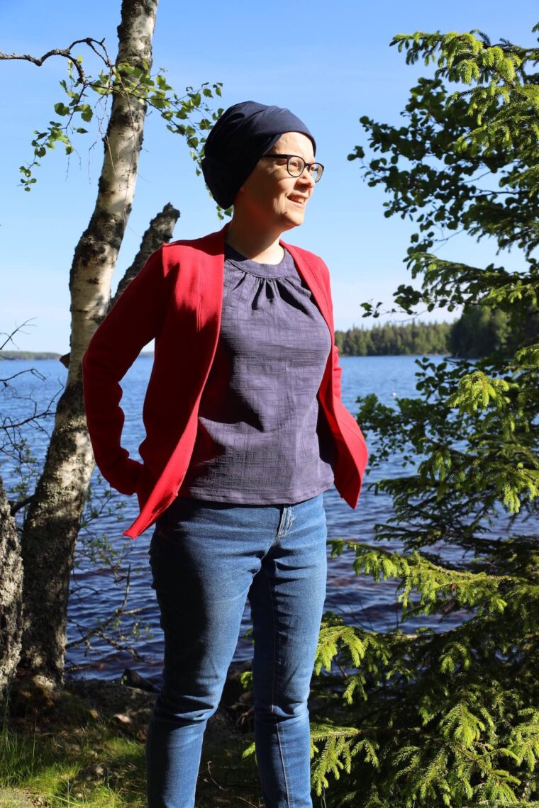 Woman is standing outside and smiling and looking into the distance. She is wearing a navy halter top layered under a red cardigan and jeans.