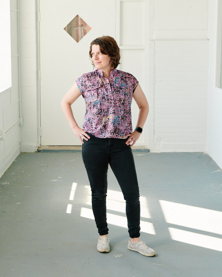 Woman standing in hallway wearing a pink Santa Rosa Top and black jeans. She is looking out a window.