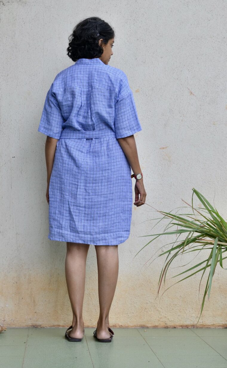 Back view of a woman wearing blue linen Santa Rosa Dress. The dress is belted She is standing outside in front of a white wall.