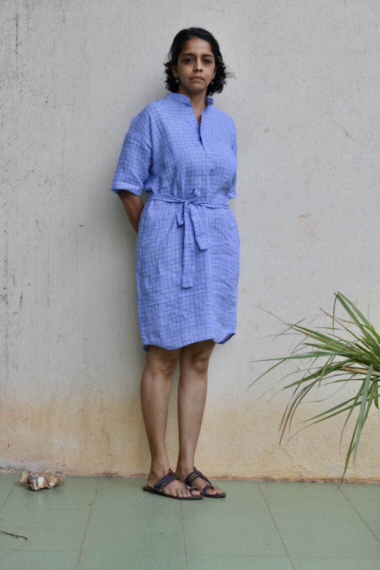 Woman wearing blue linen Santa Rosa Dress. The dress is belted She is standing outside in front of a white wall.
