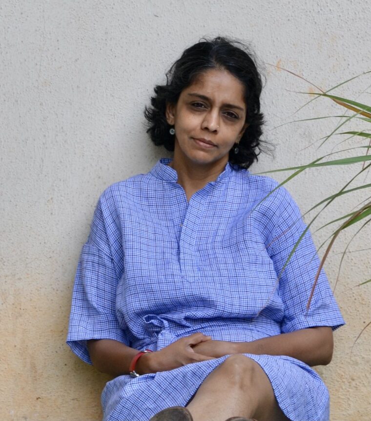 Woman sitting and smiling. She is looking directly at the camera. She is wearing a blue checked linen fabric. The dress is belted at the waist