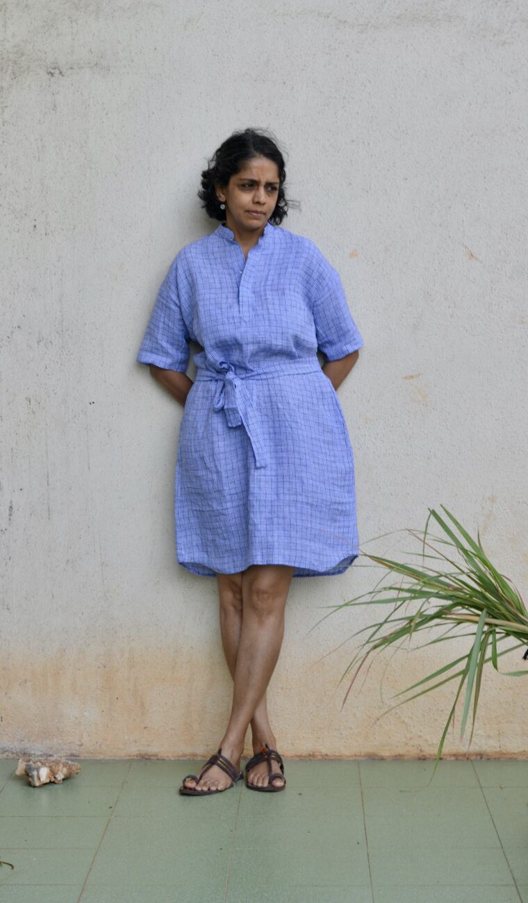 Woman wearing blue linen Santa Rosa Dress. The dress is belted She is standing outside in front of a white wall.