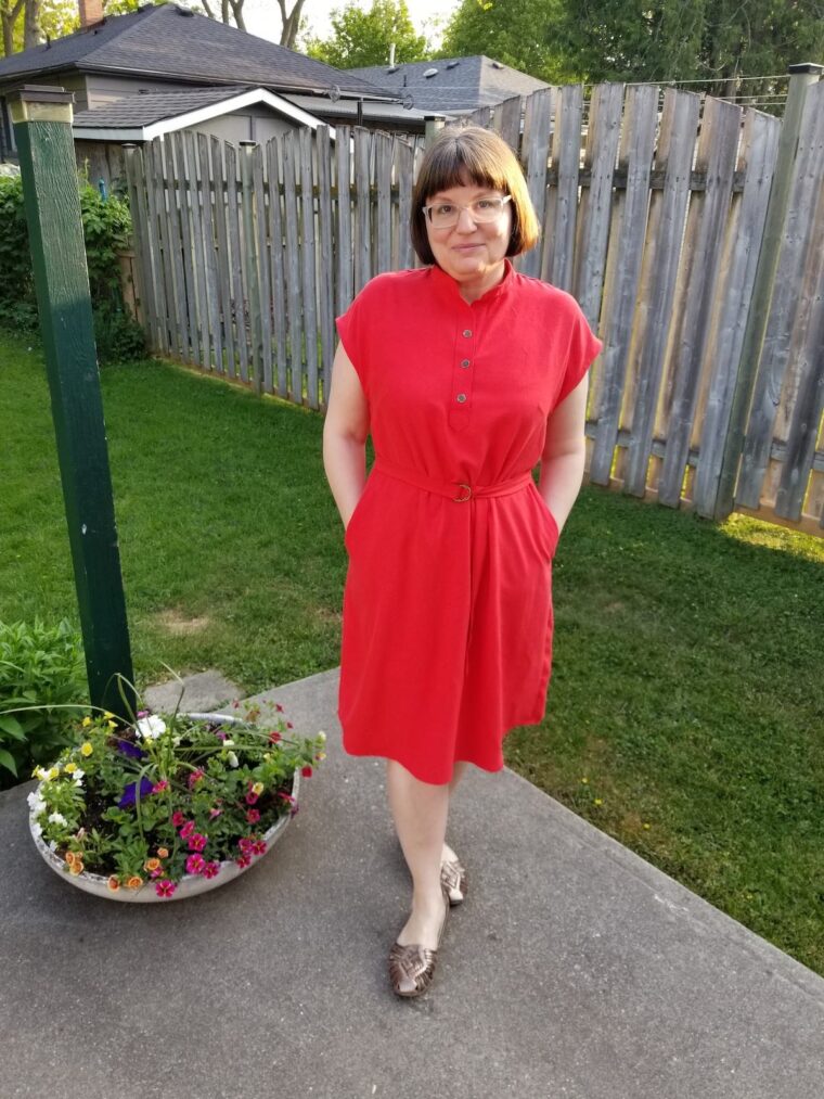 Woman standing in a backyard and smiling, wearing a red short sleeve pullover dress with a button placket at the neckline. She has a belt made in the same fabric.