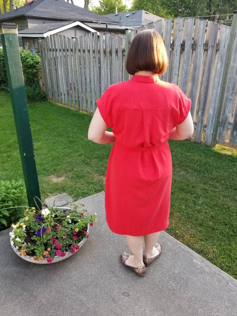 Back view of a woman standing in a backyard and smiling, wearing a red short sleeve pullover dress with a button placket at the neckline. She has a belt made in the same fabric.