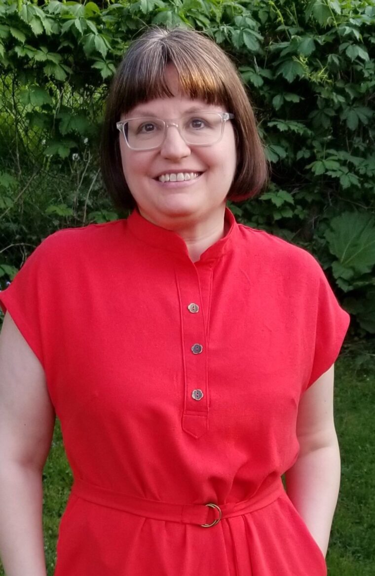 Close up of a woman standing in a backyard and smiling, wearing a red short sleeve pullover dress with a button placket at the neckline. She has a belt made in the same fabric.