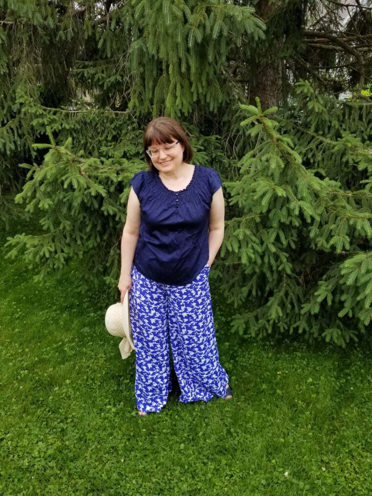 Woman wearing wide-legged trousers with a navy shirt and holding a straw sunhat.