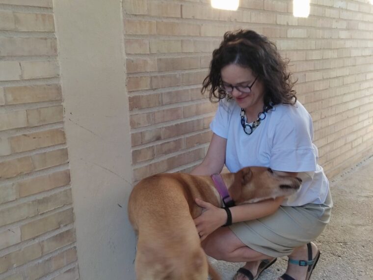 Woman in white t-shirt and khaki skirt, squatting down to pet a tan colored dog.