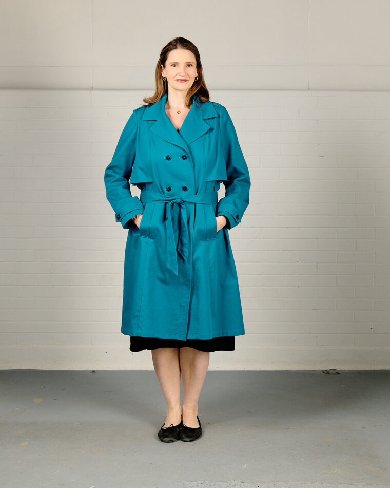 Woman is standing in front of a white brick wall wearing a bright blue trench coat that is buttoned and belted.